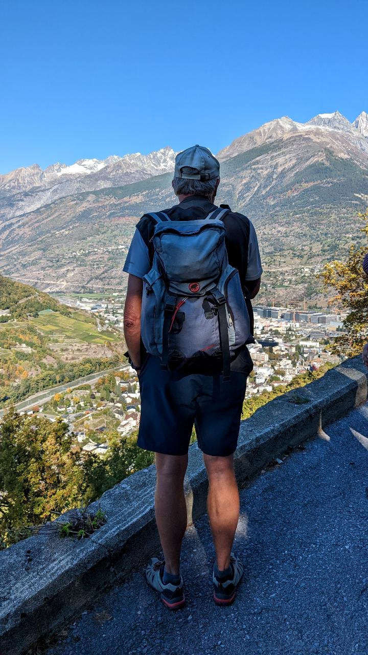 Anselmo can only be seen from behind. He carries a backpack and looks down into the valley to Visp. In the background are mountains and a blue sky.