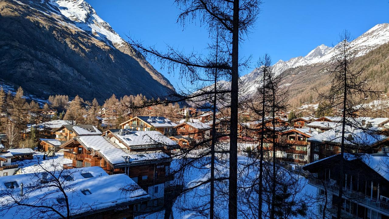 Evening atmosphere. Snowy houses of Winkelmatten Zermatt. In the foreground fir trees and in the background mountains with snow and a blue sky.