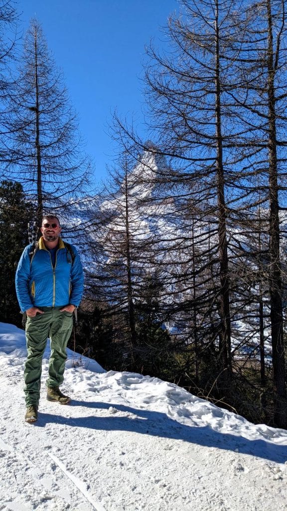 Der Boden ist schneebedeckt. Bäume säumen den Wegrand und bilden einen Wald. Zwischen den Zeigen kann man das Matterhorn sehen. Der Himmel ist stahlblau. Matthias posiert auf der linken Bildhälfte.