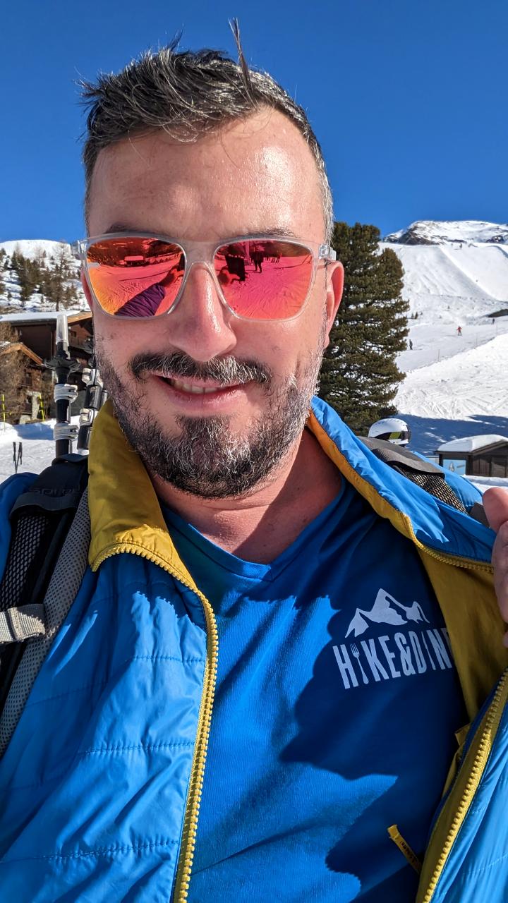 The photo is a selfie. Matthias took a picture of himself posing with sunglasses, a smile and the blue Hike&amp;Dine T-shirt. In the background, you can see the Riffelalp in the snow and the blue sky.