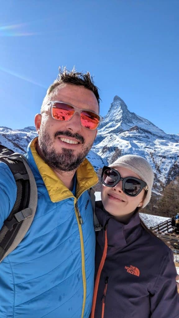 Solène and Matthias take a selfie with the Matterhorn. Both wear sunglasses and smile at the camera. A few rays of sunlight graze the left side of the picture. The sky is blue.