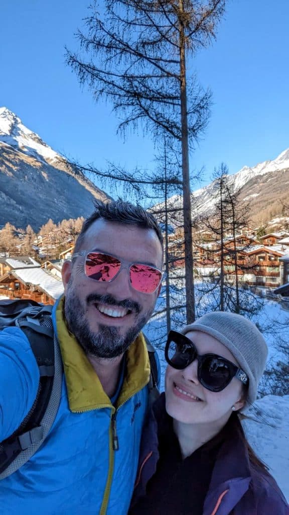Matthias and Solène, both wearing sunglasses, pose for a selfie. In the background, you can see blue sky, snowy mountains, and Valais houses.