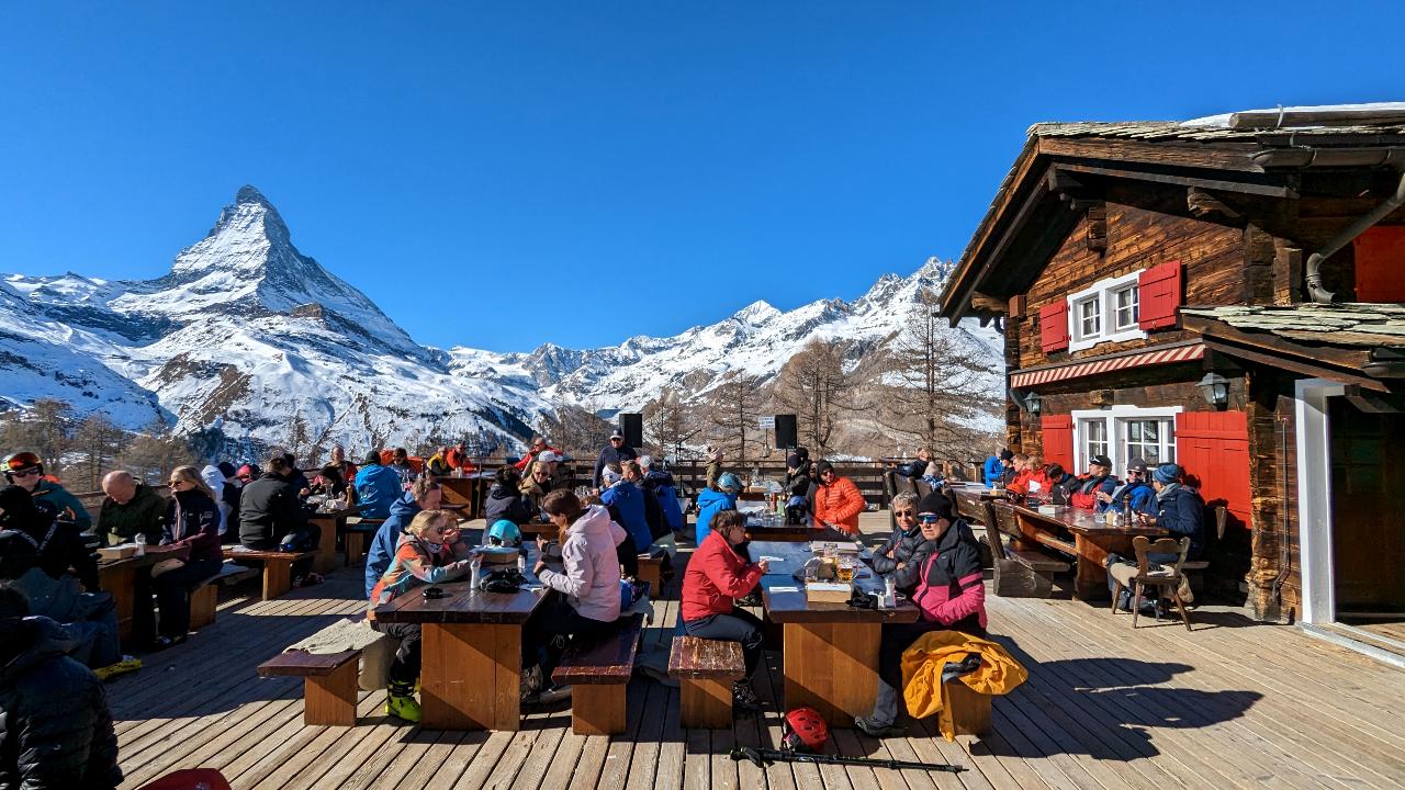 Mountain restaurant Alphitta with snow-free but sun-full terrace. Floor, tables, and benches are made of wood. Many people enjoy the sun and food. In the background, the Matterhorn.
