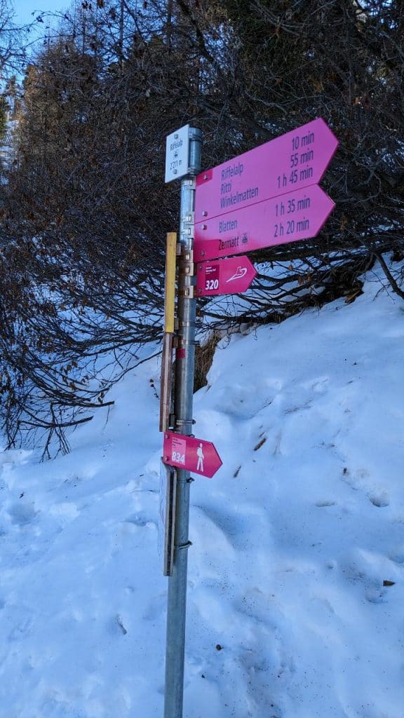 Ein pinkfarbener Wegweiser mit der Beschriftung Riffelalp, Ritti, Winkelmatten, Blatten, Zermatt. Der Wegweiser ist im Schnee.