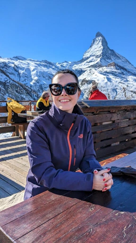 Solène sits at a wooden table on the Riffelalp, has her hands folded and smiles at the camera. In the background, you can see the Matterhorn and the clear blue sky.