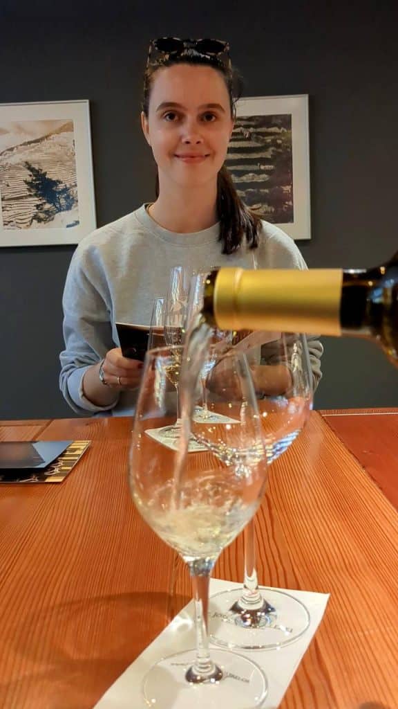 Solène hält eine Weinbroschüre in der Hand. Vor Solène wird ein Glas Weisswein eingeschenkt-