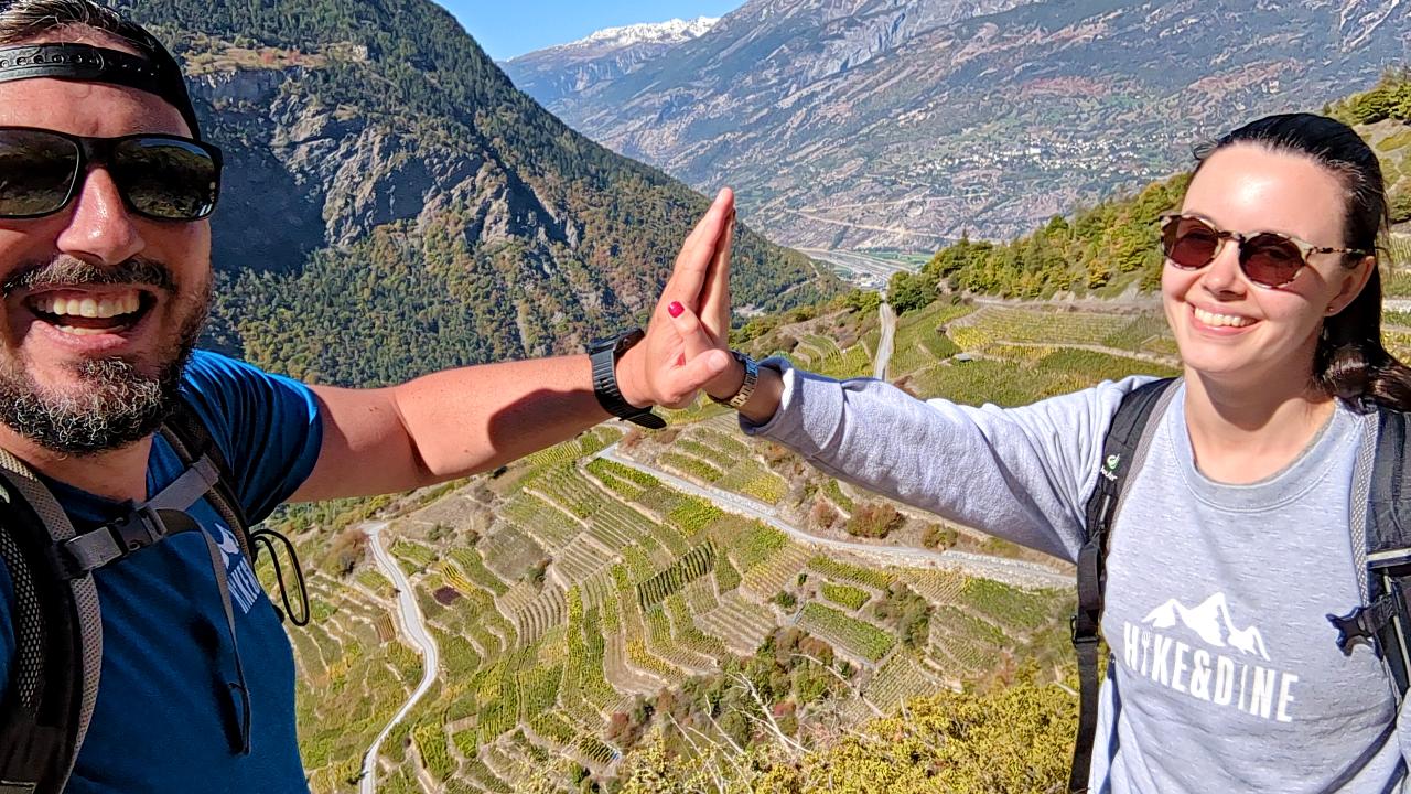 Matthias and Solène shake hands. In the background, you can see the vineyard of St. Jodern Kellerei. Behind the vineyard, other mountains of Valais can be seen.