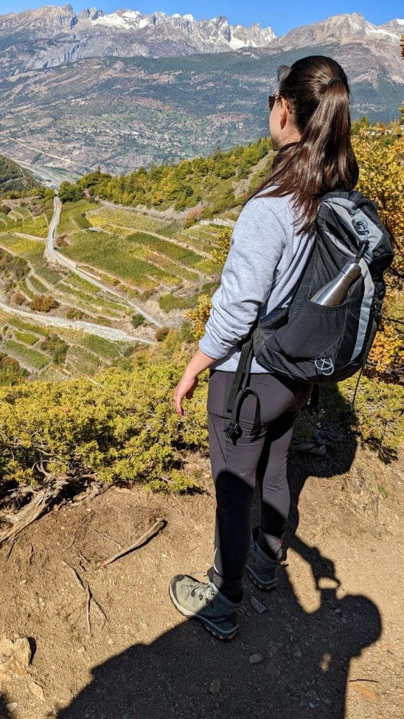 Solène looks down at the vineyard. She is carrying a backpack. In the background, one can see mountains.