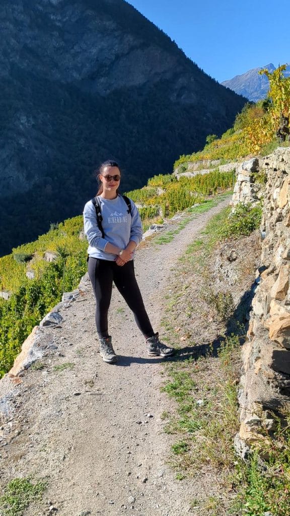 Solène carrying sunglasses and the Hike&amp;Dine shirt, smiling at the camera. The hiking trail leads downwards on the Reblehrpfad towards Visp.