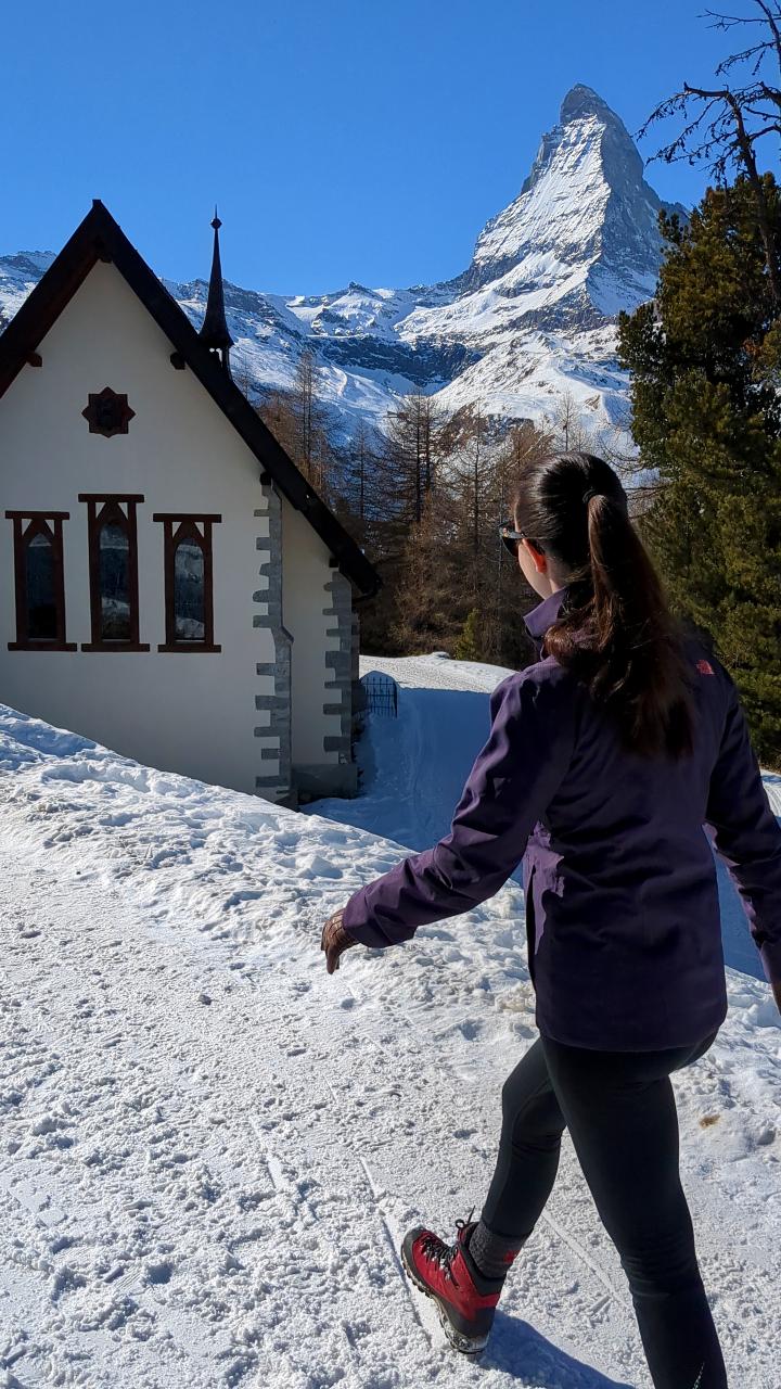 Soléne walks on a winter hiking trail. The snow on the path is rolled. On the left side, you can see a small chapel. In the background, the Matterhorn is enthroned. The sky is steel blue.