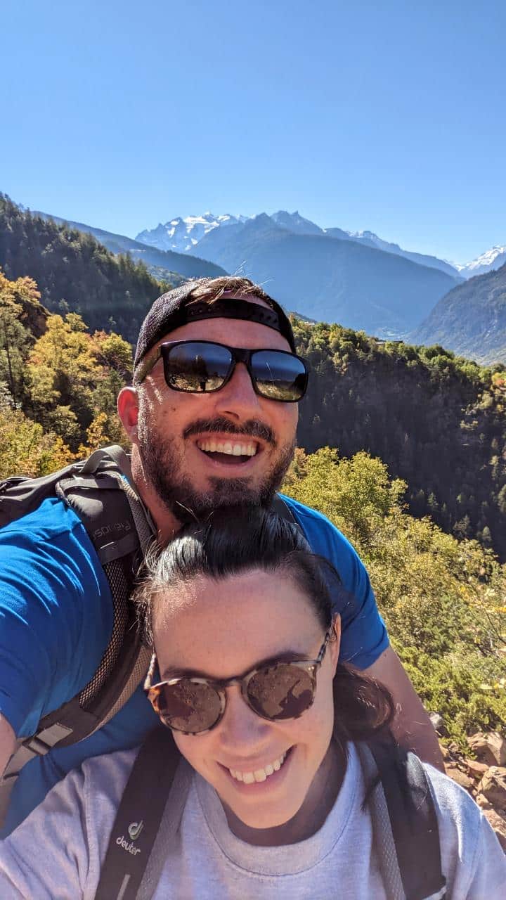 Solène and Matthias take a selfie and laugh happily into the camera. In the background the forest and the mountains Kleine Furggen, Dürrenhorn, and Platthorn.