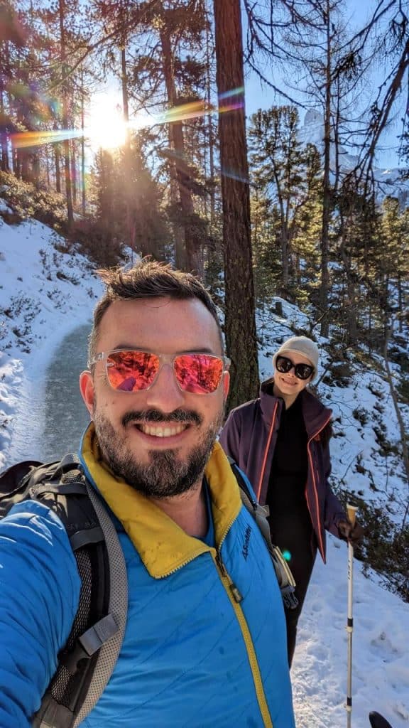 Matthias und Solène sind auf dem Selfie zu sehen. Sie lachen in die Kamera. Im Hintergrund sieht man den präparierten Winterwanderweg und die untergehende Sonne, die durch die Bäume hindurchscheint.