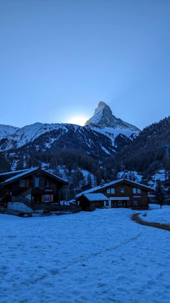 View of Matterhorn from Schluhmattstrasse. The sun sets behind the Matterhorn. The sky is clear. In the foreground, there are houses and a meadow, both covered with snow.