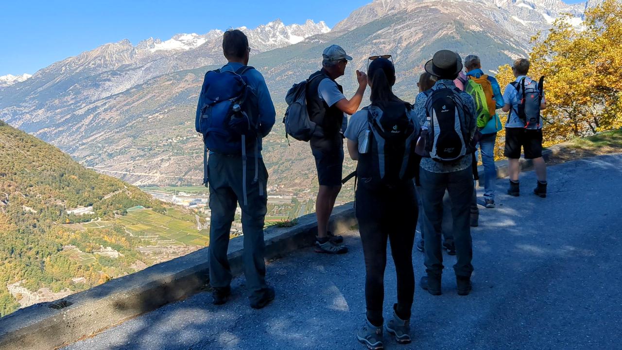 Several people (Imbach hiking group) looking at the mountain panorama and listening to the hiking leader telling stories. All of them can only be seen with their backs.