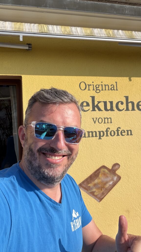 Matthias de Hike&Dine se tient tout sourire devant l'écriteau « Original Gersauer Käsekuchen vom Dampfofen » au restaurant Traube à Gersau.