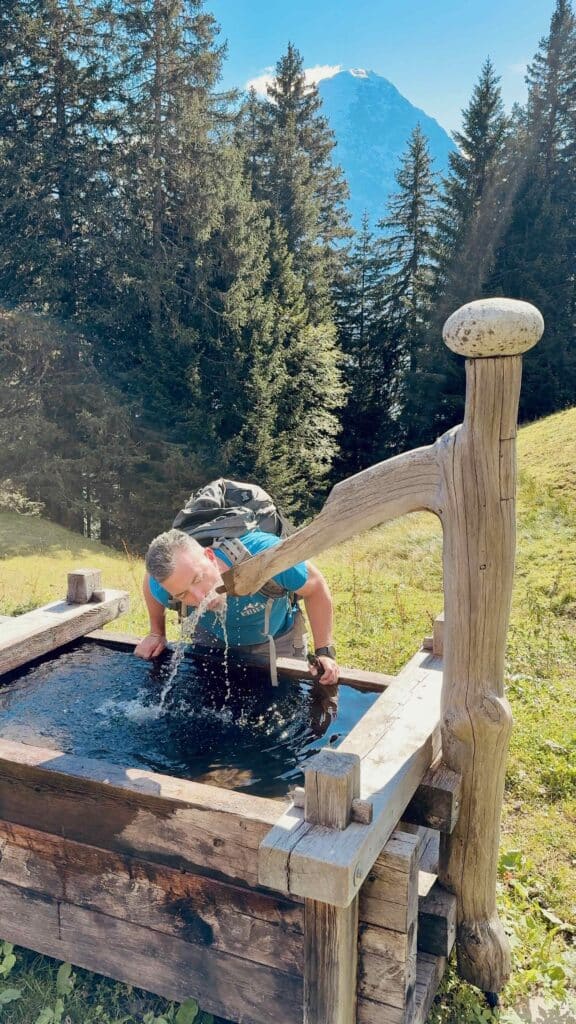 Matthias erfrischt sich an einem rustikalen Holzbrunnen mit frischem Bergwasser auf dem Weg zur Bussalp.